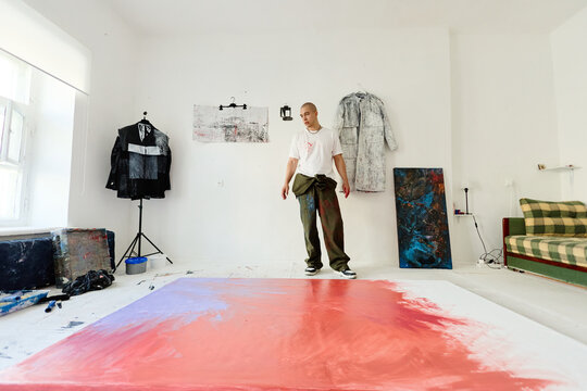 Wide shot of young adult man creating contemporary artwork in studio, standing near large canvas and painted pieces. Useful for creative industry marketing, artist branding, exhibition promotion