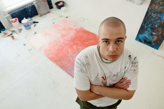 Portrait of young adult Caucasian male artist standing with crossed arms in studio beside abstract canvas, presenting creative profession and branding use for art business, high angle