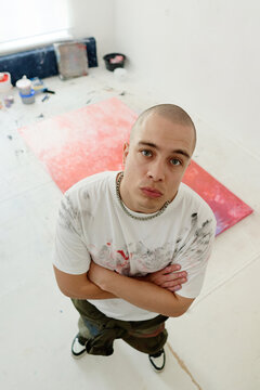 Vertical portrait of young adult Caucasian male artist standing with folded arms in studio beside canvas, presenting contemporary art identity. Useful for creative branding, gallery promotion