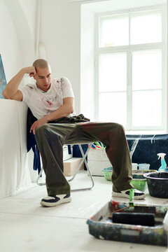 Vertical shot of young adult Caucasian man sitting in art studio, resting and reflecting after painting session. Suitable for creative profession, artist lifestyle, burnout concepts