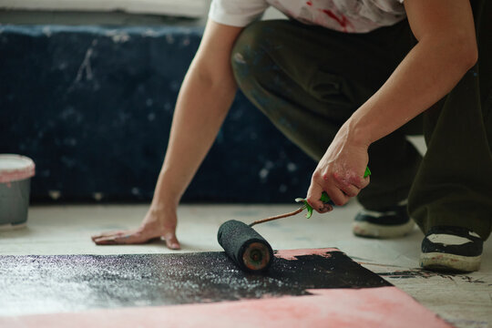 Close up of young adult artist painting large canvas on studio floor with roller, creating contemporary artwork. Ideal for creative industry branding, art education, editorial use