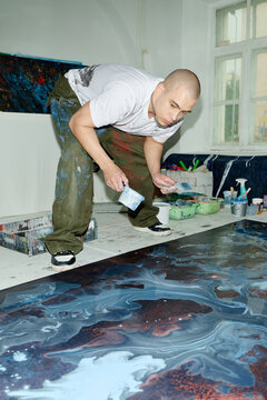 Vertical shot of young adult Caucasian male artist bending and spreading paint across large canvas in studio, creating contemporary artwork. Useful for creative industry marketing, artist branding