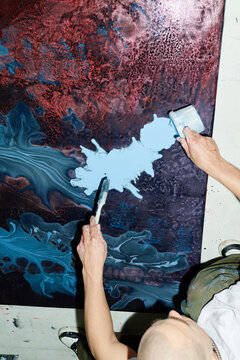 Vertical shot of contemporary artist spreading paint across abstract canvas with paintbrush and bucket, creating textured artwork. Ideal for creative process, studio practice, modern art marketing