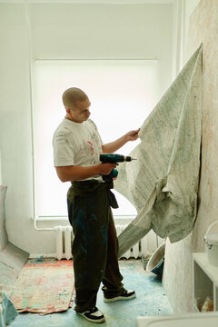 Vertical shot of young adult Caucasian man drilling and adjusting large textured canvas on in studio, creating contemporary installation. Ideal for artist branding, creative industry promotion