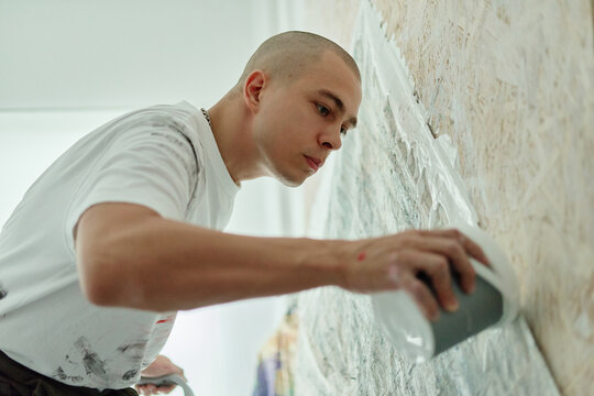 Young adult Caucasian male artist applying textured material on large canvas wall, creating contemporary artwork. Ideal for creative industry branding, studio promotion, artistic process