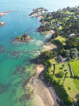 Aerial view of the Cote de Granit Rose coastline in Tregastel, featuring turquoise waters, rocky outcrops, lush green vegetation, and stone houses in Tregastel, Bretagne, France.