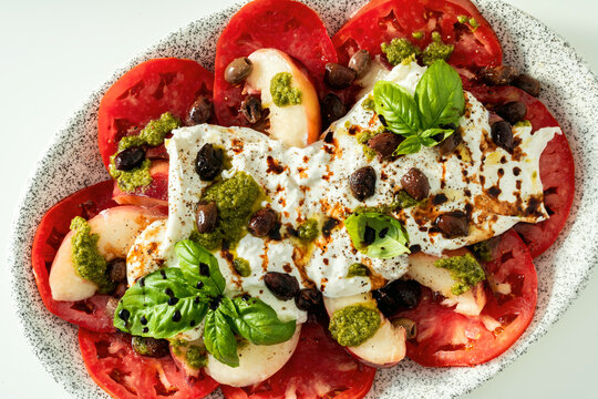 Close up of Italian style salad with tomatoes, peaches, olives, burrata and pesto on white background
