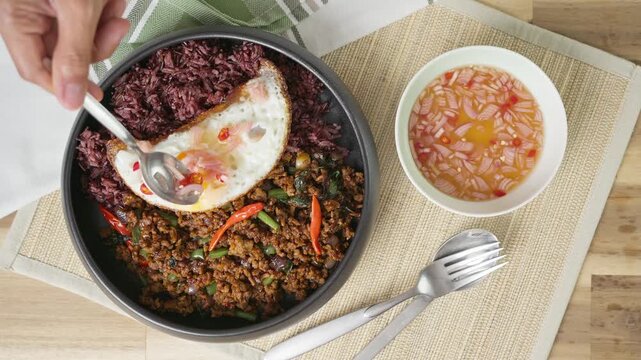 Overhead view of authentic Thai Pad Kra Pao Moo featuring spicy stir-fried minced pork with holy basil, served over Riceberry and Jasmine rice with a fried egg. Action shot of spooning Prik Nam Pla ch