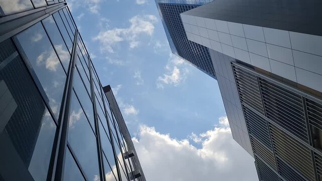 Low angle view of modern glass skyscrapers reaching toward a bright blue sky with scattered clouds in a contemporary business district.