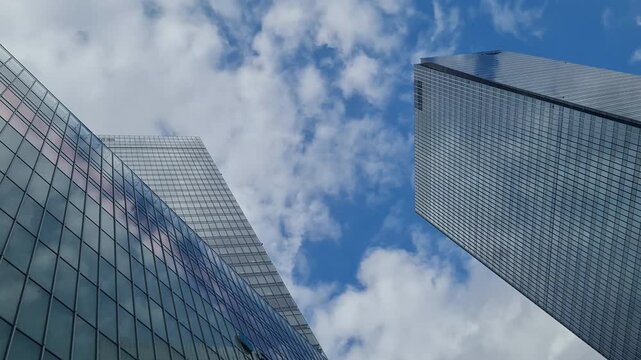 Low angle view of modern glass skyscrapers reaching towards a bright blue sky with white fluffy clouds.