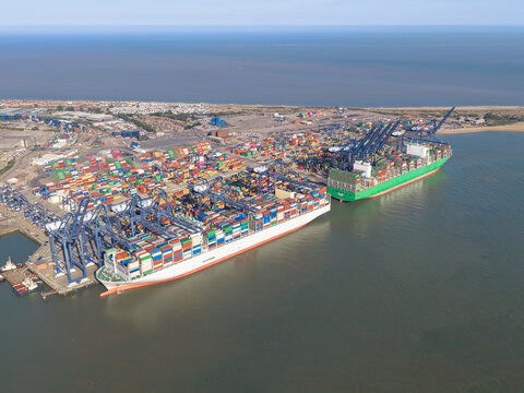 Aerial view of large container ships docked at a busy port with colorful shipping containers, industrial cranes, and harbor facilities in the United Kingdom.