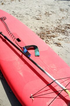 Red stand-up paddle board with paddle and coiled leash resting on sandy beach ready for summer water sports