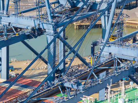 Aerial view of large blue container cranes at a busy shipping port with a cargo ship deck and calm water in the background in the United Kingdom.