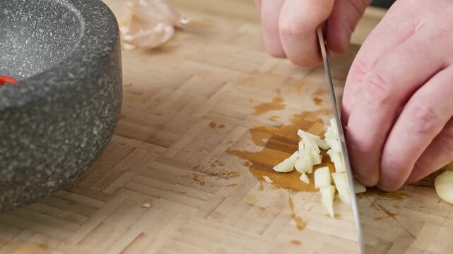 Roughly chopping fresh garlic cloves with a sharp chef knife on a wooden cutting board. Real-time food preparation footage showcasing precise slicing technique and fresh raw ingredients in a kitchen
