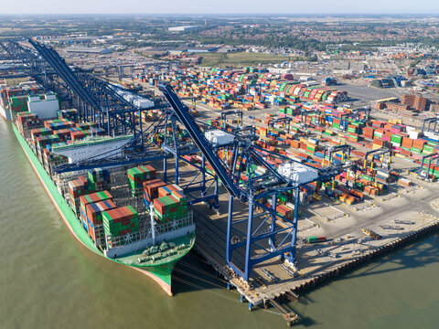 Aerial view of a large container ship docked at a busy port terminal with colorful shipping containers stacked in the yard and massive gantry cranes loading cargo in the United Kingdom.