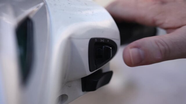 Close up of male thumb switching between high and low beam headlight on white scooter handlebar. Detail of rider hand pressing toggle switch button on motorcycle control panel, transportation concept.
