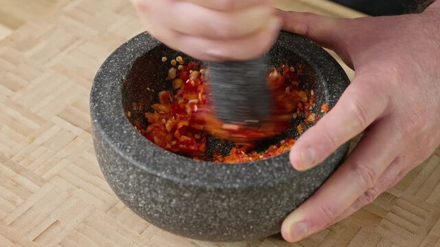 Pounding red chili peppers and fresh garlic cloves in a traditional stone mortar and pestle. Real-time action footage of crushing aromatic spicy ingredients into a fresh paste.