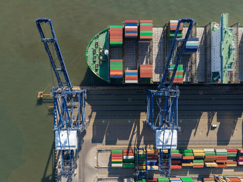 Aerial view of a large container ship being loaded with colorful shipping containers by massive blue gantry cranes at a busy port in the United Kingdom.