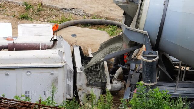 Concrete mixer truck discharges liquid cement down the chute into a pump hopper. Heavy-duty hoses supply material directly to the pipeline for pouring building foundations.