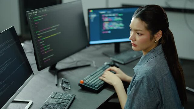Focused Young Asian female software developer typing on a mechanical keyboard, writing code on multiple computer monitors in a modern tech office. Professional IT programmer working on cyber security