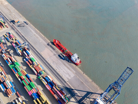 Aerial view of a red cargo ship docked at a busy port terminal with rows of colorful shipping containers and a large blue crane in the United Kingdom.