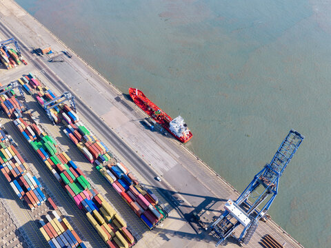 Aerial view of a red cargo ship docked at a busy shipping port with rows of colorful shipping containers and a large crane in the United Kingdom.