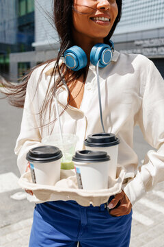 Business woman carrying coffee drinks outdoors with headphones
