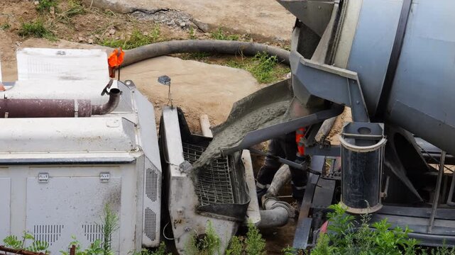 Concrete mixer truck discharges liquid cement down the chute into a pump hopper. Heavy-duty hoses supply material directly to the pipeline for pouring building foundations.