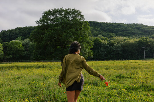 Woman walks in a field holding a flower as trees stand nearby