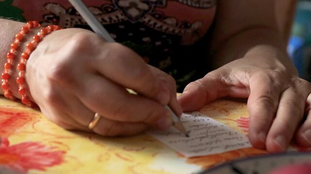 Closeup of hands writing a shopping list with a pencil on a small paper note at a kitchen table.