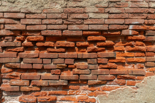 ​Horizontal photo of an ancient damaged red brick wall with eroded mortar and peeling plaster texture, showing urban decay and historic architectural detail
