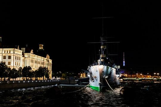 Saint Petersburg, Russia. Historic ship docked at night with illuminated waterfront buildings and city skyline in the background, reflecting on the water surface