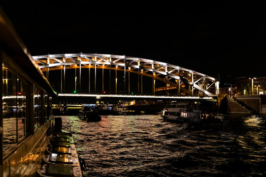 Boat night cruise concept. Illuminated bridge spans over water, with boats docked along the shore. City lights reflect on the water's surface, creating a vibrant urban scene with a tranquil atmosphere