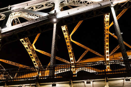 Illuminated steel bridge structure captured at night, highlighting intricate design and engineering details with glowing elements and shadows creating a striking visual impact in the urban landscape