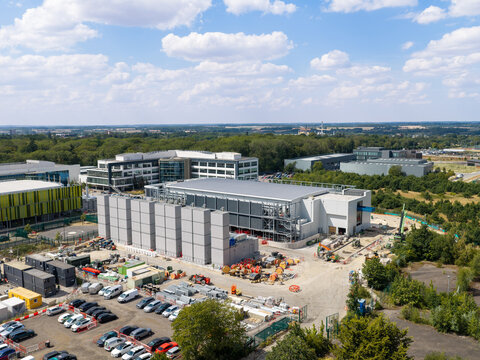 Aerial view of a large data center complex with modular cooling units, construction equipment, and a parking lot surrounded by trees in the United Kingdom.
