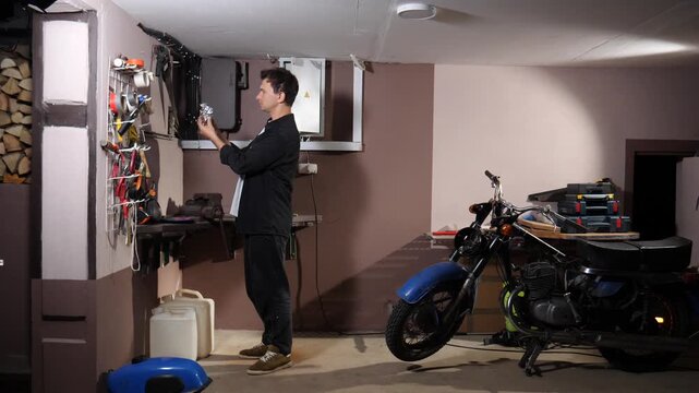 Man holding a small metallic part, standing in his workshop next to an old blue motorcycle, having tools organized on a wall rack and workbench for hobby maintenance