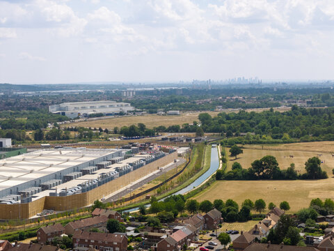 Aerial view of a large data center facility situated next to a canal with surrounding residential houses, green fields, and a distant city skyline in the United Kingdom.