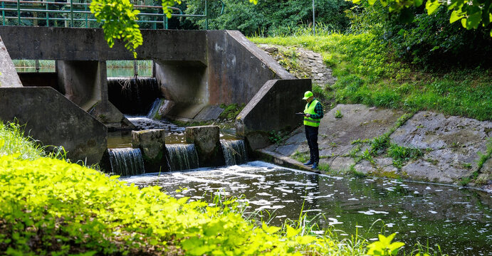 Hydraulic engineer checking water flow levels and hydro power dam equipment with digital tablet outdoors. Environmental engineer in safety vest inspecting water dam status using  plant in forest.