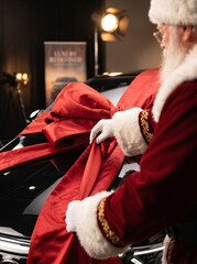 Obraz premium Santa Claus putting a large red bow on a luxury black car as a Christmas surprise gift in a professional showroom