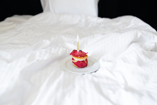 Small gourmet cake or pastry with a single lit candle on a plate, placed on a white blanket in bed. Cozy morning birthday celebration or surprise breakfast concept with selective focus.