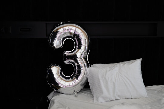 Large silver metallic balloon in the shape of number three lying on a bed with white pillows and blanket. Minimalist birthday decoration or anniversary concept, bedroom interior.