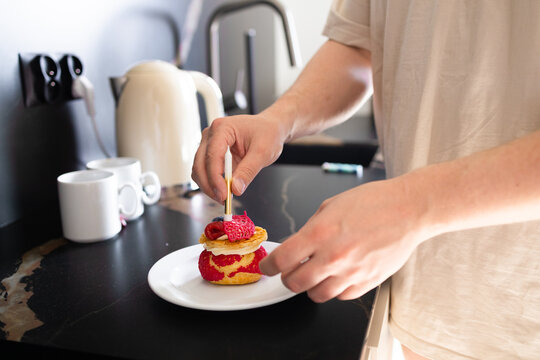 Close-up of a person's hands placing a stick or wick into a small gourmet cake with berries on a white plate. Preparing a food surprise or baking at home kitchen interior, lifestyle concept.