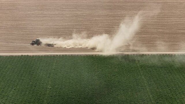 Farm tractor plowing dry agricultural field, creating huge dust cloud next to a green crop plot