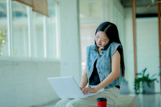 Young Asian woman reading a book with headphones and laptop by the window