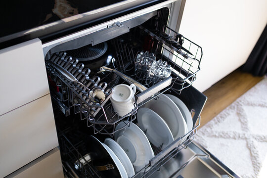 Close-up of an open modern built-in dishwasher packed with plates, mugs, and cutlery. Domestic chores, home appliance, and kitchen interior concept with selective focus.