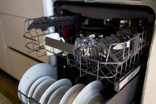 Close-up of an open modern built-in dishwasher packed with plates, mugs, and cutlery. Domestic chores, home appliance, and kitchen interior concept with selective focus.