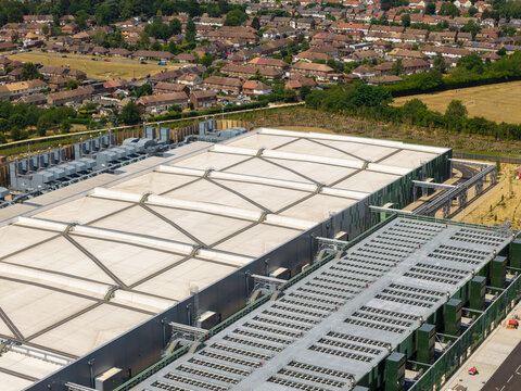 Aerial view of a large data center facility featuring industrial cooling units on its roof, situated near a residential neighborhood and open fields in United Kingdom.