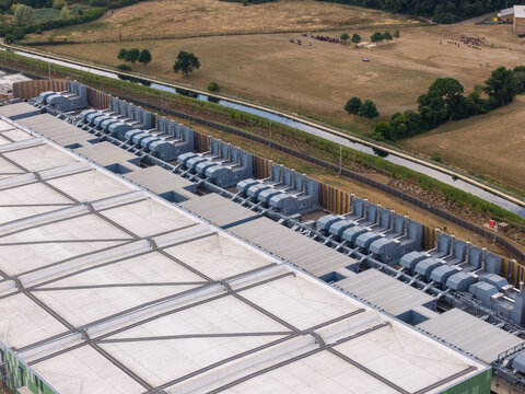 Aerial view of a large data center rooftop featuring numerous industrial cooling units and HVAC systems, situated next to a rural landscape with a canal and green fields in the United Kingdom.