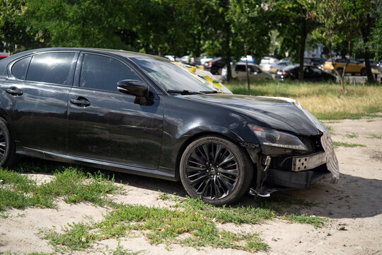 Kyiv, Ukraine. July 30, 2026. Wrecked black Lexus sedan with severe front damage parked on dirt lot with missing bumper.