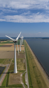 Aerial view of a row of wind turbines standing along a dike next to agricultural fields and water in Nagele, Flevoland, Netherlands.